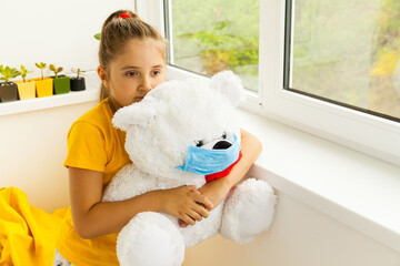 A girl and a toy bear in medical masks near the window