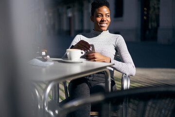 Fototapeta premium Crop cheerful young black woman browsing smartphone at table in street cafeteria