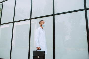 A young caucasian doctor in uniform with a protective mask on his face and a black bag walks in front of the clinic. Protection against COVID - 19 coronavirus