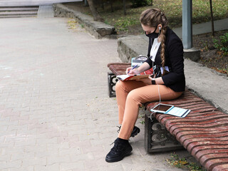 Fototapeta premium Young woman in suit sits on bench in desolate place. She listens to something on the headphones from the tablet and makes notes in notebook. Remote work or training during quarantine, with copy space