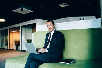 Serious caucasian male entrepreneur in formal outfit sitting with laptop computer typing mail on working process, confident businessman making research for project looking at camera in coworking space