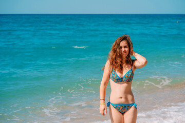 A young woman in a bright swimsuit and long hair walks on the sand along the sea with waves, clear azure water