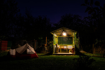 Tents versus shelter. Camping spot with white red tent and mini shelter house in the campground surounded by nature at night. No person.