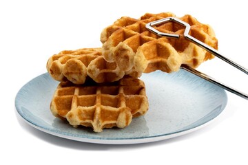 a pair of tongs placing another Belgian waffle onto a blue plate of waffles isolated on white
