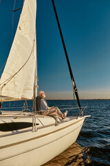 Vertical shot of carefree retired man enjoying amazing view and relaxing while sitting on the side of his sailboat or yacht deck floating in the calm blue sea