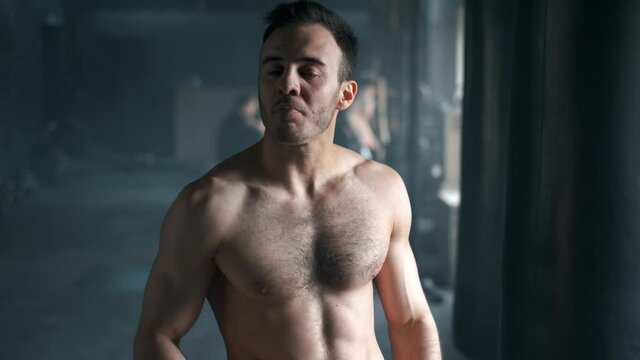 Attractive athletic shirtless young man drinking protein shake from shaker in gym and looking at camera and smiling