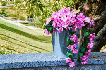 Fototapeta na wymiar Metal bucket with pink flowers on a granite parapet against the backdrop of trees and a park. Outdoor cafe decor element. Space for text. Selective focus and blurred background