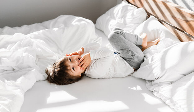 Cute little happy child boy playing in parents bed having fun at home at sunny morning. Light and shadows
