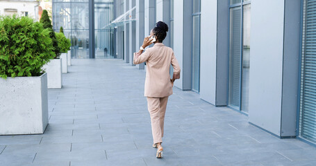 Rear of African American beautiful woman in medical mask talking on mobile phone and walking...