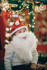Cute little boy in a false beard of Santa Claus near the New Year tree in Christmas decorations