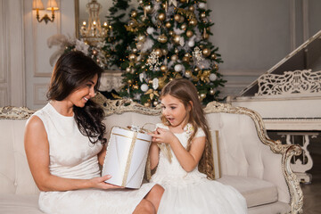 Merry Christmas and Happy Holidays. Cheerful mom and her cute daughter girl exchanging gifts in white classic interior against the background of a piano and a decorated Christmas tree. New Year 2021.