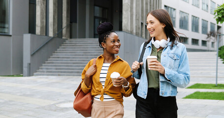 Mixed-races young pretty females best friends talking cheerfully and walking with cups of coffee...