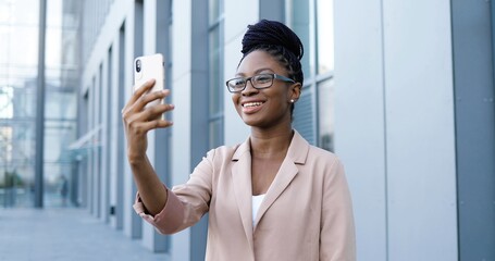 Pretty happy African American young woman in glasses having videochat on smartphone outdoor at business building. Joyful beautiful female talking and videochatting via webcam on mobile phone.