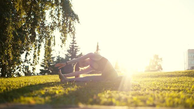 Flexible young woman sitting on yoga mat and making deflections to side, stretching outside in city park background of bright sunray and green grass in sunny day. Tracking shot in slow motion.