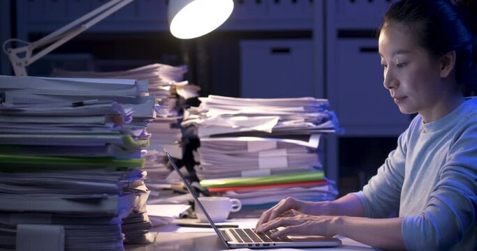 Asian business woman is sitting at desk covered with stack of paperwork and looking at paper report, typing on notebook. Alone girl checking document while working hard at late night. Side view shot.