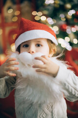 Fototapeta na wymiar Cute little boy in a false beard of Santa Claus near the New Year tree in Christmas decorations