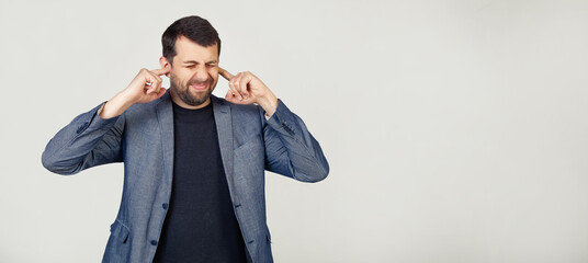 Young businessman man with a beard in a jacket covering his ears with his fingers with an annoyed expression on his face due to the noise of loud music. Deaf concept. gray background