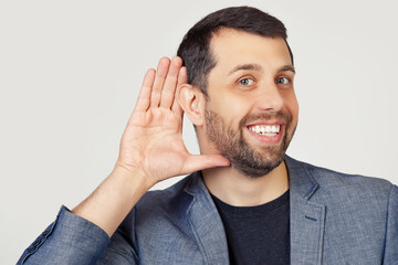 Young businessman man with a beard in a jacket, smiling with his hand above his ear, listening to rumors or gossip. Deafness concept. Portrait of a man on a gray background