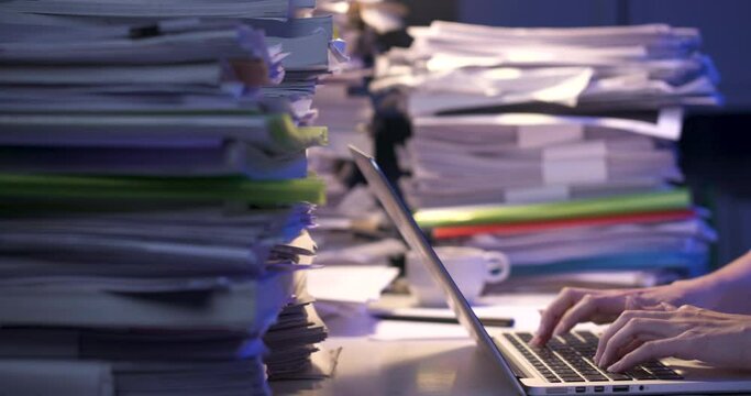 Asian business woman is sitting at desk covered with stack of paperwork and looking at paper report, typing on notebook. Alone girl checking document while working hard at late night. Close up shot.