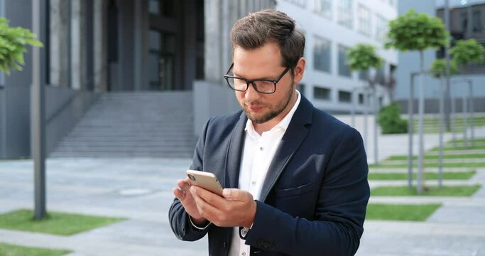 Portrait of Caucasian cheerful young stylish man tapping or scrolling on smartphone and looking at camera at street. Handsome happy male texting message on phone and smiling. Outside. Businessman.