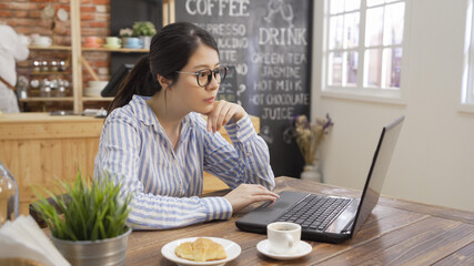 Businesswoman use laptop at modern cafe bar. Young beautiful girl freelance sitting in cozy coffee shop and working on notebook computer with cup of tea and croissant on table. waitress in background