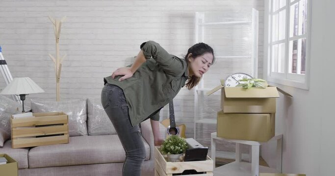 slow movement of young asian korean woman holding sore painful back while carry heavy carton container in living room moving house to new apartment. girl injured waist during relocation lifting box