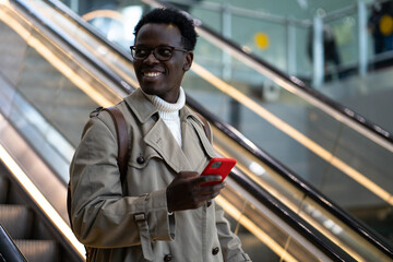 Smiling Afro-American traveler man stands on escalator in airport terminal or railway station, using mobile phone, looking aside, arrives from abroad. 