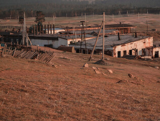 Ruins of old destroyed white factory building in field, steppe with dry yellow, red grass, electric poles, forest behind. Olkhon, sunset