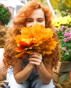 Autumn time. Ginger curly attractive girl with opened eyes wearing trendy autum color yellow shirt and blue jeans holding yellow bouquet of maple leaves near face. Enjoy autumn season. 