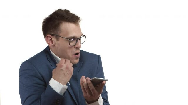 Video of funny blond guy, male entrepreneur in blue suit and glasses mumbling something at speakerphone, holding mobile phone and record voice message, standing over white background