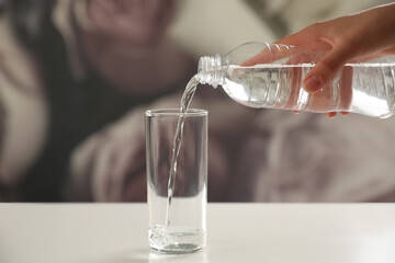 Woman pouring water from bottle into glass on table against blurred background, closeup