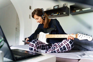Adult caucasian woman sitting on bed with earphones on head using mobile phone to play music or make a call while holding guitar in her bedroom at home - real people leisure weekend activities concept