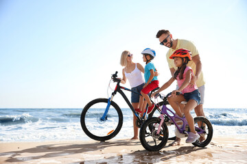 Happy parents teaching children to ride bicycles on sandy beach near sea