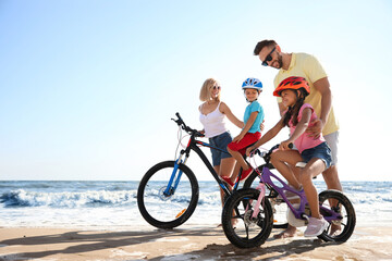 Obraz premium Happy parents teaching children to ride bicycles on sandy beach near sea