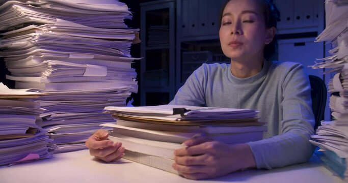 Exhausted asian business woman putting documents on desk cover with stack of paperwork. Tired alone girl looking at paper and lay down on pile of sheets while working hard at late night at home.