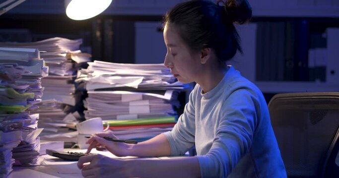 Concentrated asian business woman analyzing, calculating data, writing project reports while working hard at late night. Tired alone girl lay down on desk covered with stack of paperwork. Dolly shot