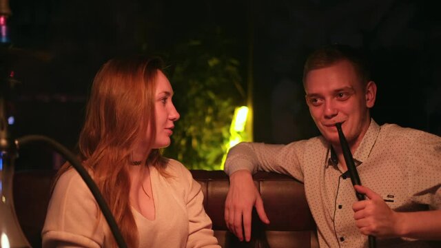 Cute couple in love relaxing on a date. Media. Caucasian young man and woman in black smoking a hookah while sitting at the table in cafe indoors together.