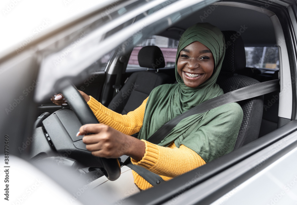 Wall mural car rent. joyful black islamic lady driving auto with fasten seat belt