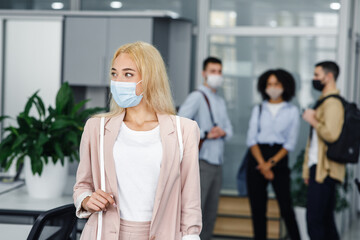 Workers in modern office hallway return to work after quarantine. Focus on beautiful blonde woman in business suit and protective mask