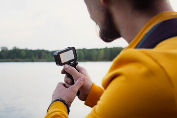 Videographer shoots a video about nature against the background of a forest and a river. A compact action camera with a white display mockup in the hands of a man.
