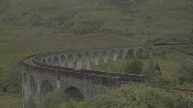 Famous steam train crossing a curve arched stone bridge in a beautiful landscape in scotland uk. Green landscape and white smoke