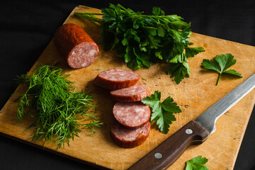 pieces of smoked sausage, parsley and dill on a cutting Board. Slicing on the light wooden cutting board with kitchen knife against the black background