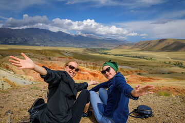 Two young women in sunglasses on hill waving arms and smiling. Female travelers are photographed against the beautiful mountains on sunny day. Active recreation and adventure.