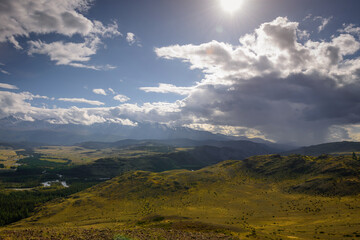 Majestic panorama of mountain plain on the background of snow-covered ridge before thunderstorm. Sun's rays break through huge clouds and beautifully illuminate green steppe and winding river.