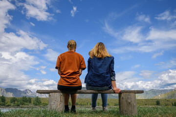 Two young women sitting on wooden bench, backs to the camera against beautiful landscape. Friends travel, admire magnificent view of mountain range under blue sky on sunny day. - Powered by Adobe