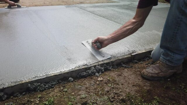Adult Caucasian male worker wearing jeans uses trowel to smooth poured grey cement, handheld pan close up