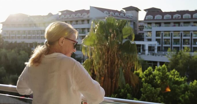 Grandmother in sunglasses stands on the balcony and waves. An old woman with wrinkles is resting on the background of a beautiful sunset or sunrise and the old city. Turkey.