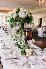 A beautiful wedding reception table set-up with tall clear glasses (trumpet vases) filled with greenery and white blooming flowers. A classic wedding floral arrangement.