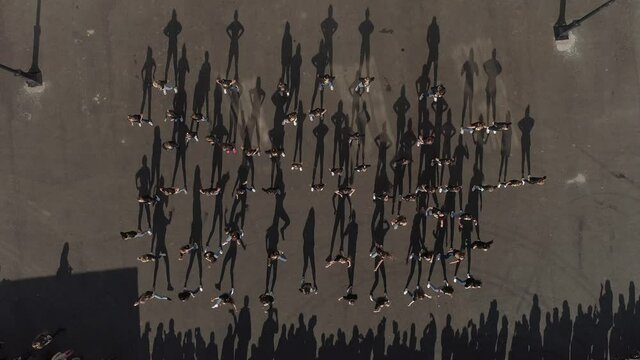 Top down aerial view of a large group of young people dancing outdoors on asphalted area. Shadow dance. A crowd of spectators nearby. Flash mob. 
