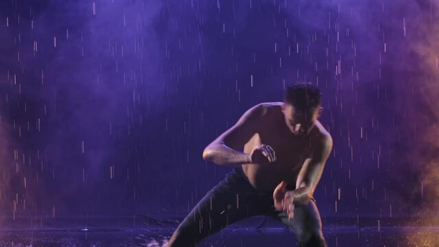A man practicing capoeira in a dark room among raindrops and splashing water. Smoky background with back light. Silhouette. Slow motion. Close up.